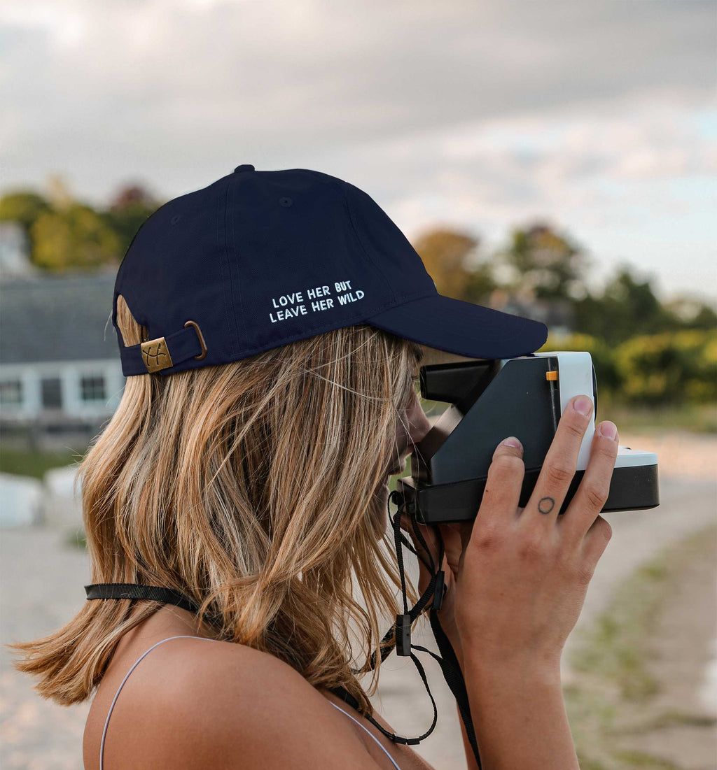 Navy blue cotton baseball cap with a white heart emblem and "love her but leave her wild" embroidered by Atticus Poetry 